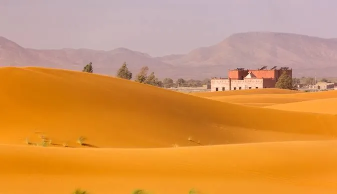 Desert landscape featuring sand dunes and a red building, illustrating the route from Tangier to Merzouga.