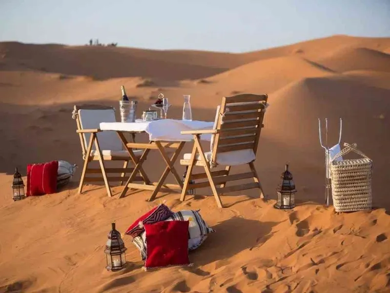 A beautifully set dinner table in the Moroccan desert, surrounded by sand dunes under a starry sky.