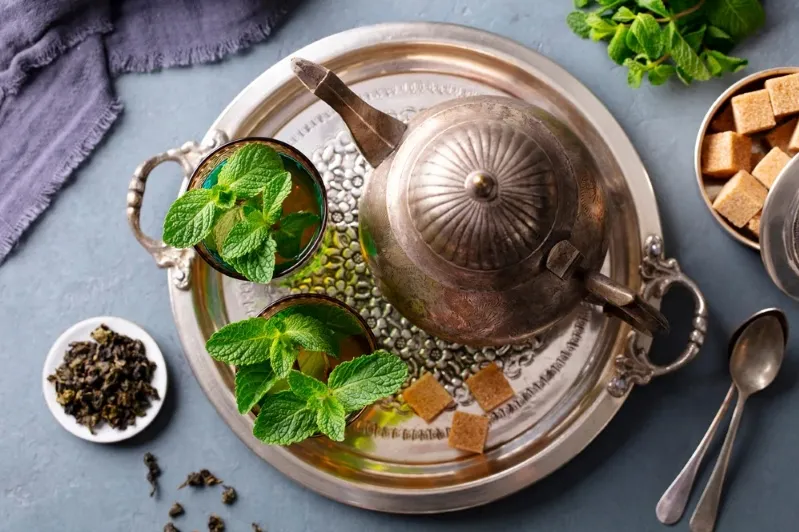A silver tea pot with fresh mint leaves beside a steaming cup of tea, highlighting the essence of Moroccan tea benefits.