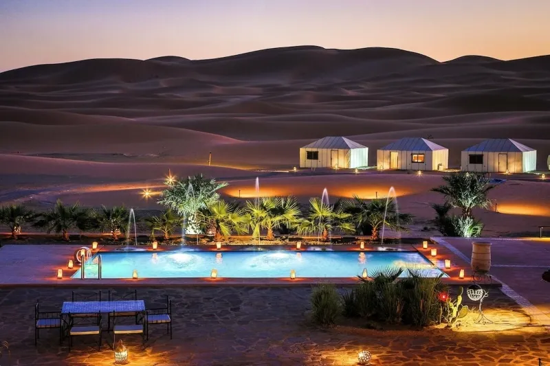 A serene desert pool at night, illuminated by lights, with a tent nearby, showcasing a desert tour marrakech  experience.