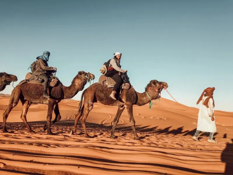 Three individuals riding camels across a vast desert landscape under a clear blue sky.