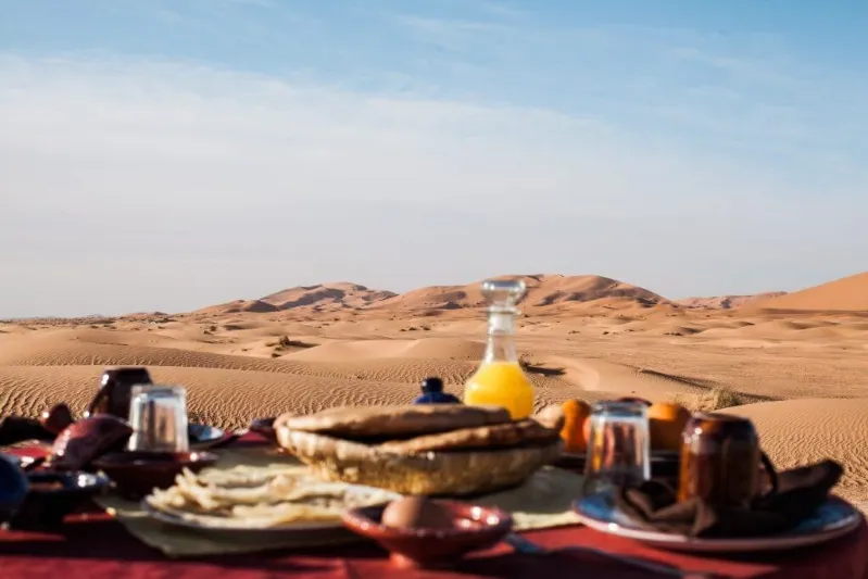 Breakfast spread in a desert setting, featuring traditional Moroccan dishes, with sand dunes in the background.