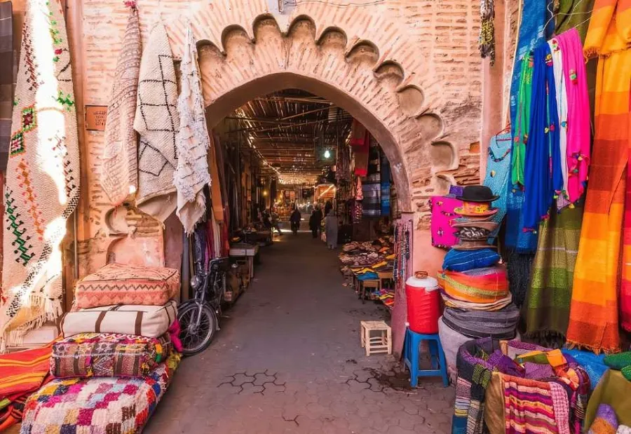 A narrow alleyway in Marrakech, Morocco, adorned with vibrant blankets and pillows for sale at a local market.