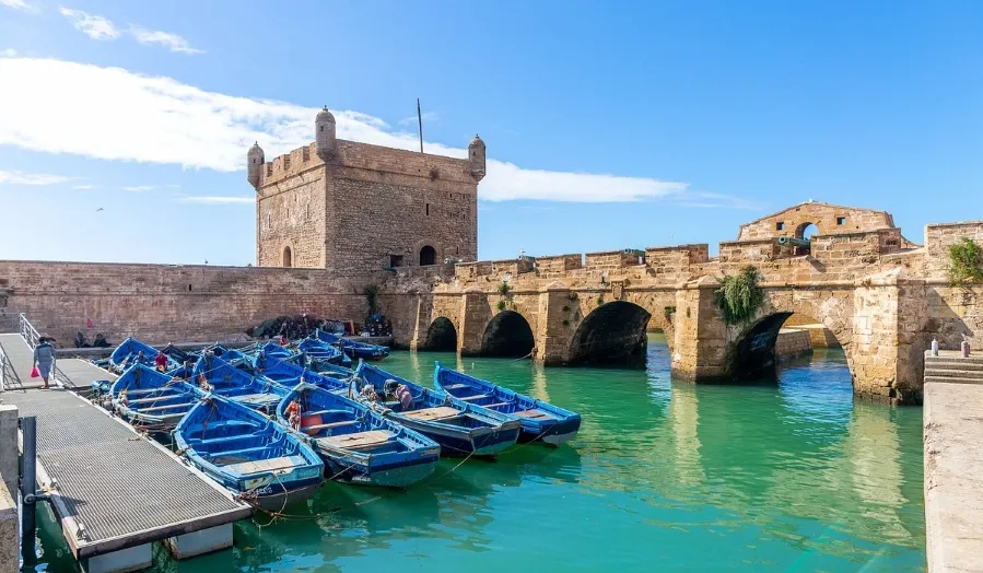 Boats are docked in the water beside a historic castle, creating a picturesque waterfront scene.
