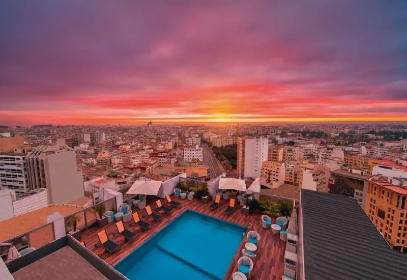 A vibrant sunset casts colorful reflections over a city pool, with the skyline of a major Moroccan city in the background.