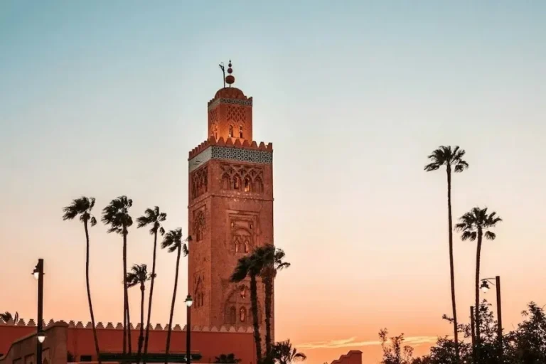 A tall, ornate minaret towers over palm trees at sunset, set against a gradient sky of orange and blue. The scene evokes tranquility and architectural beauty.