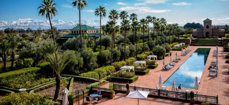 Luxurious resort scene with a long pool surrounded by palm trees and lounge chairs. Snow-capped mountains in the background under a clear blue sky. Peaceful and inviting atmosphere.