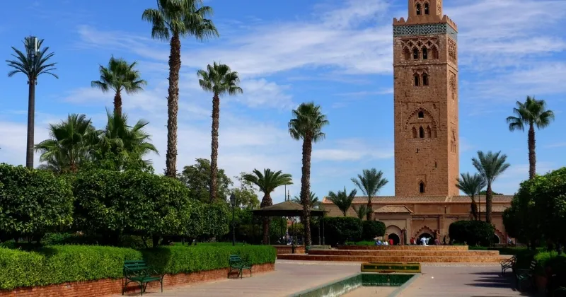A sunlit courtyard with palm trees and lush greenery surrounds a tall, ornately decorated tower. A narrow pool reflects the blue sky above.