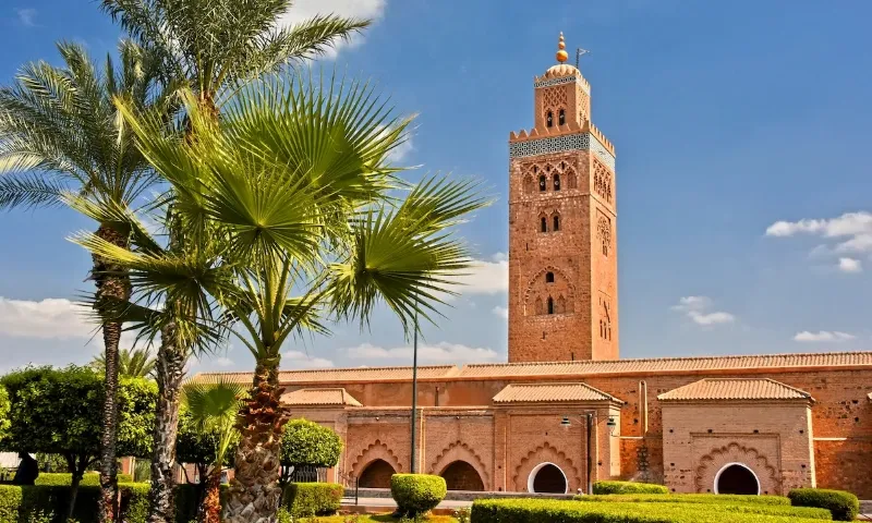Towering sandstone minaret of the Koutoubia Mosque in Marrakech, framed by palm trees and a clear blue sky, evokes a serene and historical ambiance.Between Marrakech Morocco Things to Do.